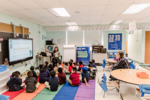 A young woman sitting in a classroom with a rainbow rug full of children listening to her read